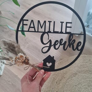 K&ouml;nnte beinhalten: Ein rundes schwarzes Wandschild mit dem Schriftzug FAMILIE Gerke in einer Kombination aus Block- und Schreibschrift. Unten befindet sich ein kleines Haussymbol mit einem Herz-Ausschnitt. Ein kleiner Strau&szlig; aus beigen und wei&szlig;en Trockenblumen ist befestigt.