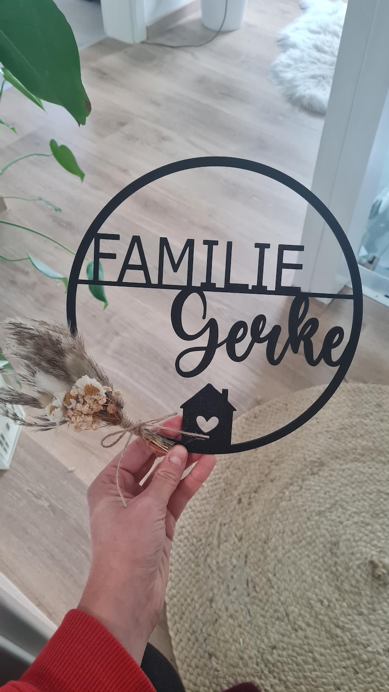 K&ouml;nnte beinhalten: Ein rundes schwarzes Wandschild mit dem Schriftzug FAMILIE Gerke in einer Kombination aus Block- und Schreibschrift. Unten befindet sich ein kleines Haussymbol mit einem Herz-Ausschnitt. Ein kleiner Strau&szlig; aus beigen und wei&szlig;en Trockenblumen ist befestigt.