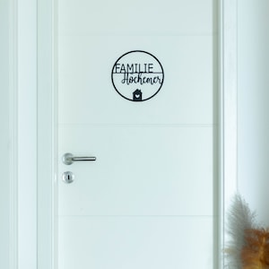 K&ouml;nnte beinhalten: Ein kreisf&ouml;rmiges schwarzes Wandschild an einer wei&szlig;en T&uuml;r. Das Schild zeigt den Text FAMILIE Hochemer in einer Kombination aus Block- und Schreibschrift sowie ein kleines Haussymbol mit einem Herz in der unteren Mitte.