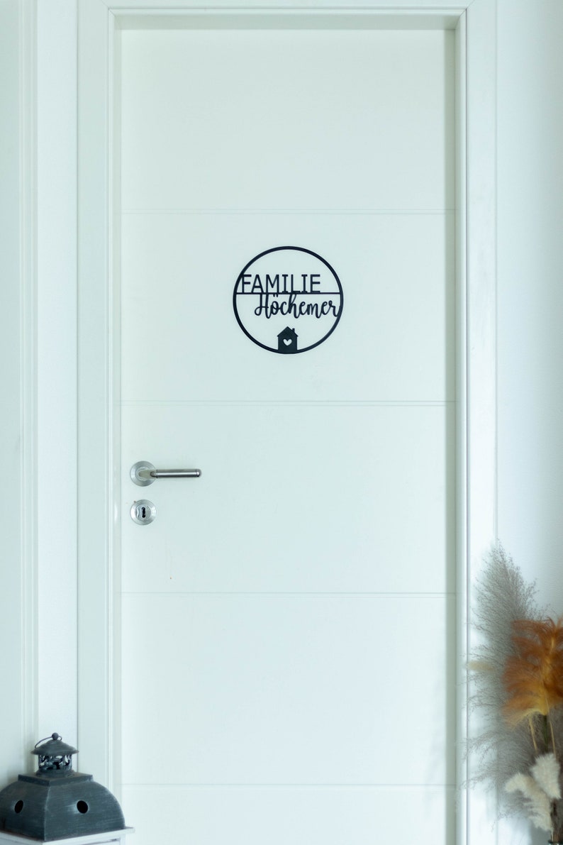 K&ouml;nnte beinhalten: Ein kreisf&ouml;rmiges schwarzes Wandschild an einer wei&szlig;en T&uuml;r. Das Schild zeigt den Text FAMILIE Hochemer in einer Kombination aus Block- und Schreibschrift sowie ein kleines Haussymbol mit einem Herz in der unteren Mitte.