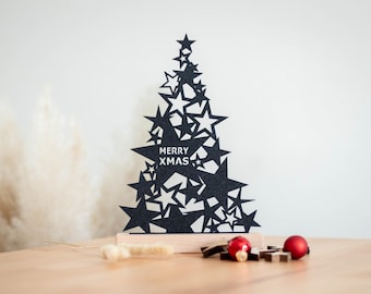 Op de afbeelding: Een zwarte glitterende kerstboom-uitsparing gemaakt van gestapelde stervormen, met de witte tekst MERRY XMAS op de centrale ster. De boom is gemonteerd op een rechthoekige, lichtgekleurde houten voet, wat een feestelijke decoratie vormt.