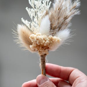 K&ouml;nnte beinhalten: Ein Ansteckbl&uuml;mchen bestehend aus einer Auswahl getrockneter botanischer Elemente, darunter wei&szlig;es farnartiges Blattwerk, flauschiges beiges und wei&szlig;es Hasenschwanzgras sowie kleine cremefarbene Bl&uuml;ten, die mit braunem Garn umwickelt sind.