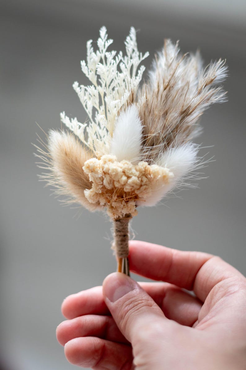 K&ouml;nnte beinhalten: Ein Ansteckbl&uuml;mchen bestehend aus einer Auswahl getrockneter botanischer Elemente, darunter wei&szlig;es farnartiges Blattwerk, flauschiges beiges und wei&szlig;es Hasenschwanzgras sowie kleine cremefarbene Bl&uuml;ten, die mit braunem Garn umwickelt sind.
