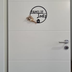 K&ouml;nnte beinhalten: Ein schwarzes, kreisf&ouml;rmiges Wandschild mit dem Text FAMILIE Lentz und einem kleinen Haussymbol mit Herz-Ausschnitt. Ein kleines B&uuml;ndel getrockneter beiger und wei&szlig;er Blumen ist an der linken Seite des kreisf&ouml;rmigen Rahmens an einer wei&szlig;en T&uuml;r befestigt.