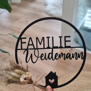 K&ouml;nnte beinhalten: Ein schwarzes, kreisf&ouml;rmiges Wandschild mit dem Schriftzug FAMILIE Weidemann in einer Kombination aus Block- und Schreibschrift. Das Design enth&auml;lt eine kleine Haussilhouette mit Herz-Ausschnitt und einen kleinen Trockenblumenstrau&szlig; mit Bindfaden.