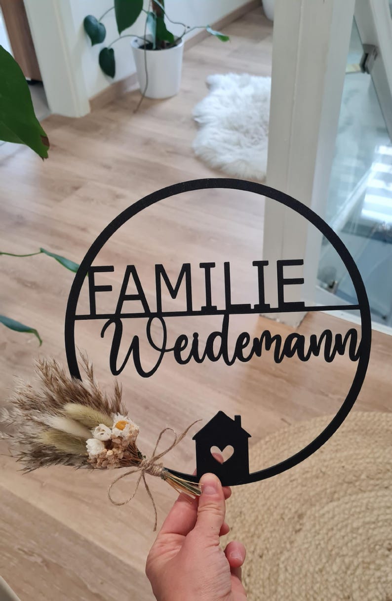 K&ouml;nnte beinhalten: Ein schwarzes, kreisf&ouml;rmiges Wandschild mit dem Schriftzug FAMILIE Weidemann in einer Kombination aus Block- und Schreibschrift. Das Design enth&auml;lt eine kleine Haussilhouette mit Herz-Ausschnitt und einen kleinen Trockenblumenstrau&szlig; mit Bindfaden.