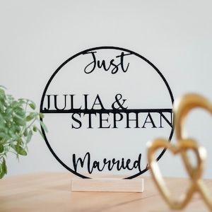 Könnte beinhalten: Ein schwarzes kreisförmiges Schild mit einer horizontalen Linie durch die Mitte, das den Text Just, JULIA & STEPHAN und Married zeigt. Das Schild ist auf einem rechteckigen hellen Holzsockel montiert und steht aufrecht.