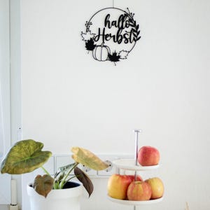 Op de afbeelding: Een zwarte cirkelvormige wanddecoratie met de tekst "hallo Herbst" in cursief schrift, versierd met silhouetten van bladeren en pompoenen. Daaronder staat een witte etag&egrave;re met rode en gele appels en een witte plantenpot.