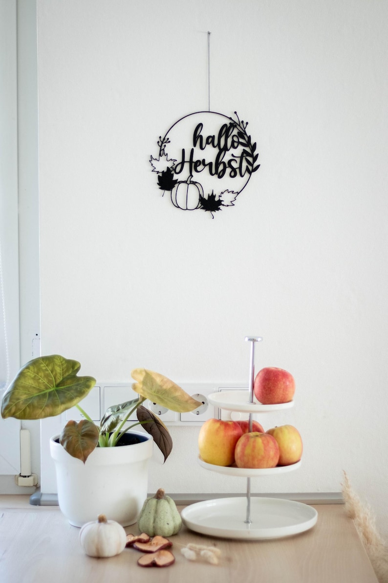 Op de afbeelding: Een zwarte cirkelvormige wanddecoratie met de tekst "hallo Herbst" in cursief schrift, versierd met silhouetten van bladeren en pompoenen. Daaronder staat een witte etag&egrave;re met rode en gele appels en een witte plantenpot.