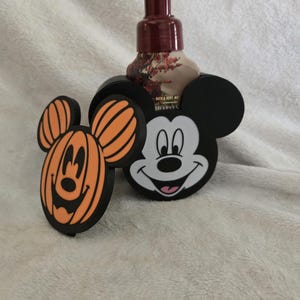 Könnte beinhalten: Ein dunkelroter Seifenspender mit Pumpe steht in einem schwarzen Halter in Form von Mickey Mouse. Daneben befindet sich ein separates schwarzes und orangefarbenes Dekostück in Form eines Mickey Mouse-Kürbisses. Das Etikett zeigt Bath & Body Works.
