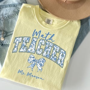 May include: A light yellow t-shirt with the words "Math TEACHER" in blue script and floral print. A blue bow graphic and the name "Ms. Manson" are also printed on the shirt. The shirt is folded on top of blue jeans and a tan hat.