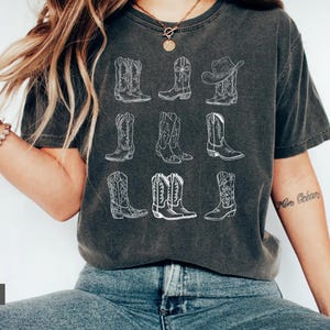 May include: Dark gray t-shirt featuring a white line drawing of nine different cowboy boots and a cowboy hat. The boots vary in style, with intricate stitching details. The shirt is worn by a person.