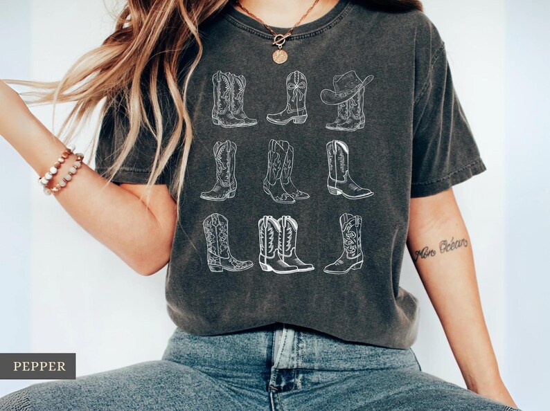May include: Dark gray t-shirt featuring a white line drawing of nine different cowboy boots and a cowboy hat. The boots vary in style, with intricate stitching details. The shirt is worn by a person.