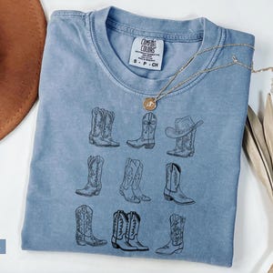 May include: Blue jean colored t-shirt with a crew neck and short sleeves. The shirt features a black line drawing of nine different cowboy boots. A gold necklace with a pendant is draped across the shirt.