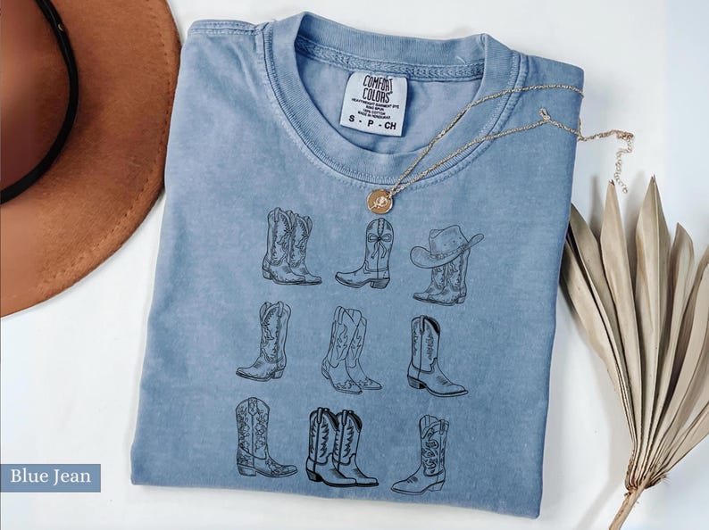 May include: Blue jean colored t-shirt with a crew neck and short sleeves. The shirt features a black line drawing of nine different cowboy boots. A gold necklace with a pendant is draped across the shirt.