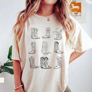May include: Ivory-colored t-shirt featuring a collection of nine different cowboy boot designs in black line art. The boots vary in style, with some having intricate patterns and others a more classic look. The word "IVORY" is printed at the bottom.