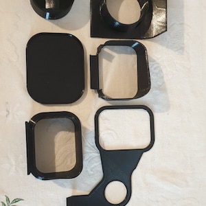 Pode incluir: Uma variedade de componentes pretos impressos em 3D, incluindo uma peça circular com uma alça, uma moldura quadrada com uma abertura circular e várias molduras retangulares e quadradas. As peças estão dispostas sobre uma toalha de mesa floral.