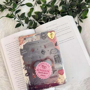 以下が含まれることがあります： 「Hopeless Romantic Readers Society」と書かれたハート型のポップソケットを含む、ロマンチックでゴシックな要素のコラージュが施された透明な携帯電話ケース。ケースには金のハートチャームと様々なイラストが描かれています。
