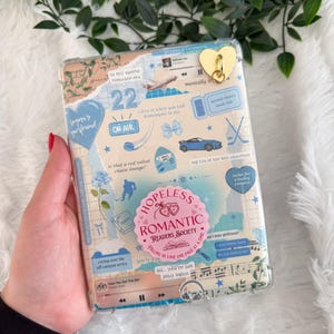 Może przedstawiać: Przezroczyste etui na telefon ozdobione niebieskimi i beżowymi naklejkami, z tekstem takim jak "Hopeless Romantic Readers Society". Dołączony jest złoty uchwyt na telefon w kształcie serca. Etui jest trzymane w dłoni.