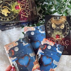 May include: Several books and e-reader covers are shown. The books have black covers with gold zodiac symbols and the title "Zodiac Academy". The e-reader covers are blue with a heart design and various text elements.