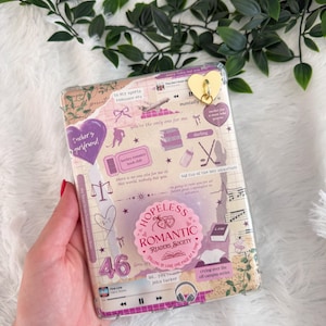 Puede incluir: Una funda transparente para teléfono con un diseño de collage con el texto "Hopeless Romantic Readers Society". La funda tiene un dije dorado en forma de corazón y varias pegatinas con frases como "Tucker's girlfriend".