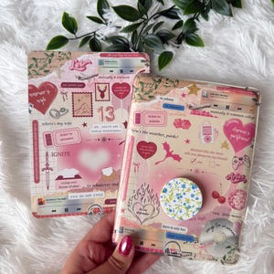 May include: Two decorative phone cases with a collage design. The cases feature a pink and beige color scheme with various graphics, including hearts, text, and a floral pop socket. One case has the word "IGNITE".