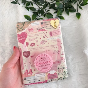 Puede incluir: Una funda transparente para teléfono con un diseño de collage con corazones rosas, notas musicales y el texto "Hopeless Romantic Readers Society". Un dije dorado en forma de corazón está adjunto en la parte superior.