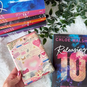 Puede incluir: Una pila de libros con cubiertas coloridas, incluyendo títulos como "Binding 13" y "Releasing 10" de Chloe Walsh. Una cubierta de libro estampada se sostiene en una mano. Los libros están sobre una superficie blanca y esponjosa con follaje verde.