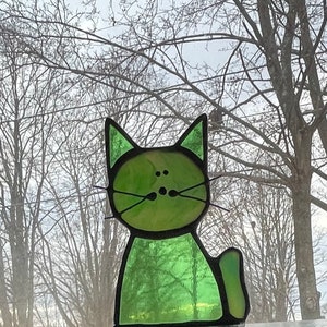 May include: A green stained glass cat figurine with black outlines. The cat is sitting with its tail curled around its body. The background is a blurry image of trees.