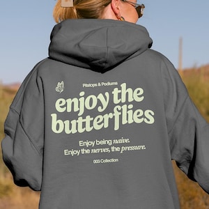 Op de afbeelding: Een donkergrijze hoodie met de tekst "enjoy the butterflies" in een lichtgroen lettertype op de achterkant. De hoodie heeft ook extra tekst eronder. De achtergrond toont een woestijnlandschap.