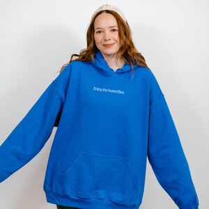 May include: A vibrant blue hoodie with a front pocket and a drawstring hood. The phrase "Enjoy the butterflies" is printed in white on the chest. The hoodie is a comfortable, casual garment.