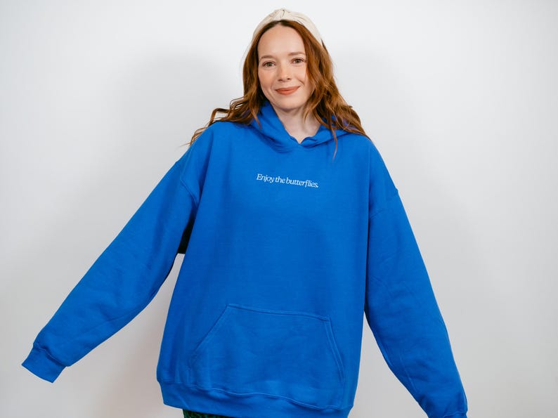 May include: A vibrant blue hoodie with a front pocket and a drawstring hood. The phrase "Enjoy the butterflies" is printed in white on the chest. The hoodie is a comfortable, casual garment.