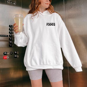 May include: White hoodie with a black print that says "F2002". The model is wearing grey shorts.