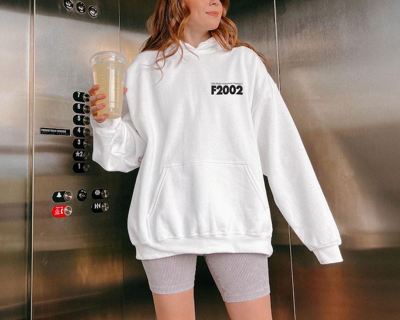 May include: White hoodie with a black print that says "F2002". The model is wearing grey shorts.