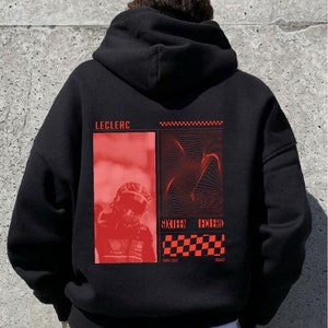 Puede incluir: Sudadera con capucha negra con un estampado gráfico rojo que presenta un piloto de carreras y el texto "Leclerc" y "Sixteen Hundred".