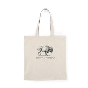 May include: A beige tote bag featuring a detailed black and white illustration of a bison. Below the bison is the text "42.8869° N, 78.8789° W". The bag has two long handles and is made of a sturdy fabric.