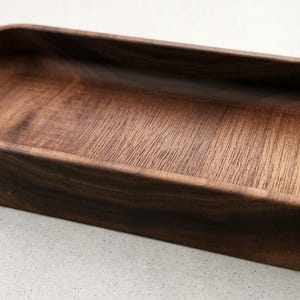 Könnte beinhalten: Ein rechteckiges Holztablett mit abgerundeten Ecken. Das Tablett besteht aus dunkelbraunem Holz mit sichtbaren Maserungsmustern. Der Innenraum ist heller als die Außenseite, was einen Kontrast erzeugt. Das Tablett ist leer und steht auf einer hellen Oberfläche.