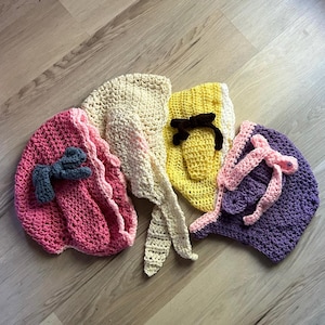 May include: Four handmade crochet bonnets in pink, cream, yellow, and purple. Each bonnet features a unique design, including bows and decorative accents. The bonnets are displayed on a wooden surface, showcasing their intricate details and vibrant colors.