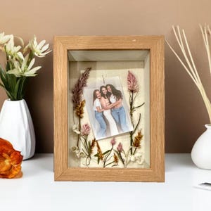 Peut inclure: Un cadre en bois avec une photo de trois personnes, entourée de fleurs et de feuillages séchés. Le cadre est rectangulaire avec une finition en bois naturel. Le fond est en tissu beige clair.