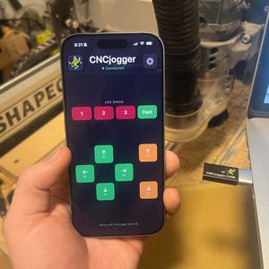 May include: A smartphone displays the "CNCjogger" app interface, showing speed controls and directional arrows. The screen is held in a hand, with a laptop and a USB drive labeled "CNCjogger.com" visible in the background.