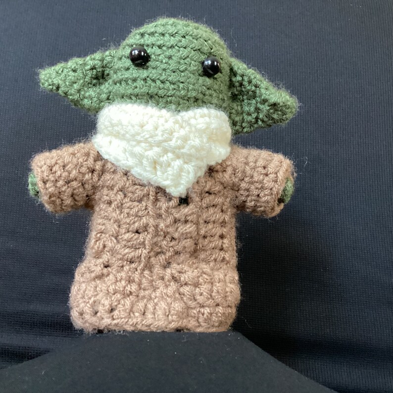 May include: A green crocheted plush toy with large black eyes wearing a brown crocheted sweater and a white crocheted scarf.