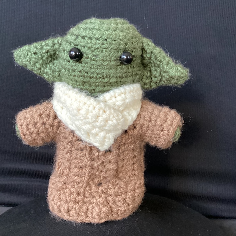 May include: A crocheted green and brown plush toy with large black eyes. The toy is wearing a white crocheted scarf.