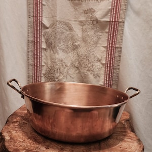 May include: A copper cooking pot with two handles sits on a wooden surface. Behind it is a linen towel with the text "LA GASTRONOMIE FRANCAISE" and a rooster illustration. The pot has a shiny, reflective surface.