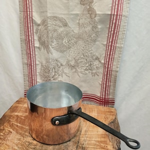 Op de afbeelding: Een ronde koperen steelpan met een gepolijste metalen binnenkant en een lange, donkere, platte ijzeren steel die is bevestigd met een enkele zilverkleurige klinknagel. De buitenkant van de pan heeft een warme, reflecterende koperen afwerking.