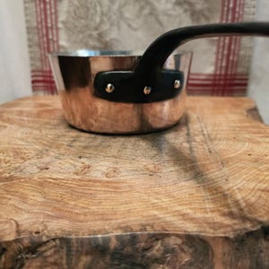 May include: A copper saucepan with a polished metallic finish and a black handle. The handle is securely attached to the side of the pan with three visible silver-toned metal rivets. The pan has a cylindrical shape with a flat base and straight sides.