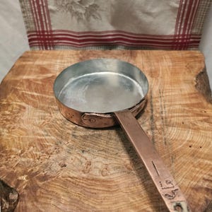 May include: A round copper pan with a silver-toned interior and a long, flat copper handle. The handle features visible markings and a small hole at the end. The exterior of the pan shows a warm, metallic copper finish with a riveted handle attachment.