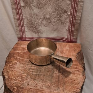 May include: A polished brass saucepan with a short, cylindrical handle attached to the side. The pan features a smooth, metallic finish and a circular opening, showcasing a simple, functional design with visible rivets connecting the handle to the body.