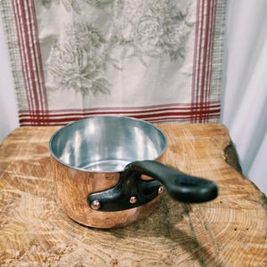 May include: A round copper saucepan with a polished silver-toned interior and a dark, heavy-duty handle attached with two copper rivets. The side of the pan features a small stamp that reads Made in France, indicating its origin.