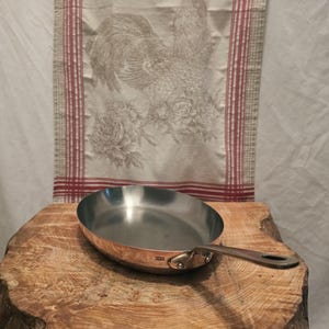 May include: A round copper frying pan with a stainless steel interior and a long metal handle. The side of the pan features a small stamp that reads Made in France. The pan has a polished metallic finish and is positioned on a wooden surface.