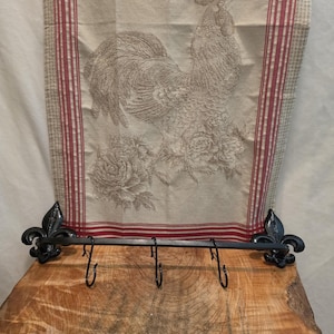 May include: A black metal wall rack with three hooks and fleur-de-lis ends, positioned in front of a beige kitchen towel featuring a rooster illustration, floral details, red striped borders, and the text LA GASTRONOMIE FRANÇAISE.
